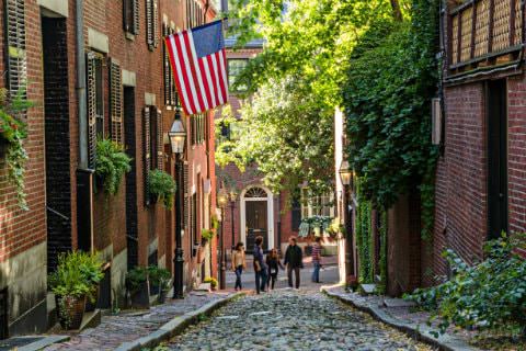 A few people at the end of a sloped stone alley between red brick buildings with green plants and vines