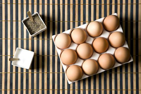 Three white containers of a dozen brown eggs, salt, and pepper on a bamboo placemat