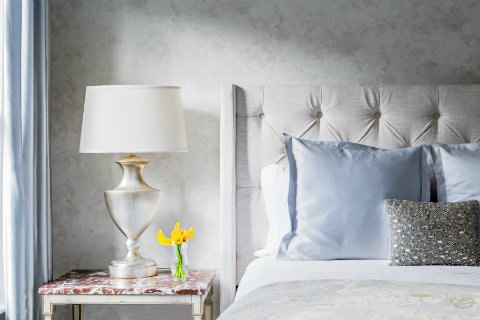 Bed with beige tufted headboard, white sheets and blue pillows, white nightstand with white-washed gold lamp and yellow flowers