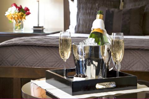 Black tray of champagne chilling in silver bucket and two filled champagne flutes with view of elegant bed in background