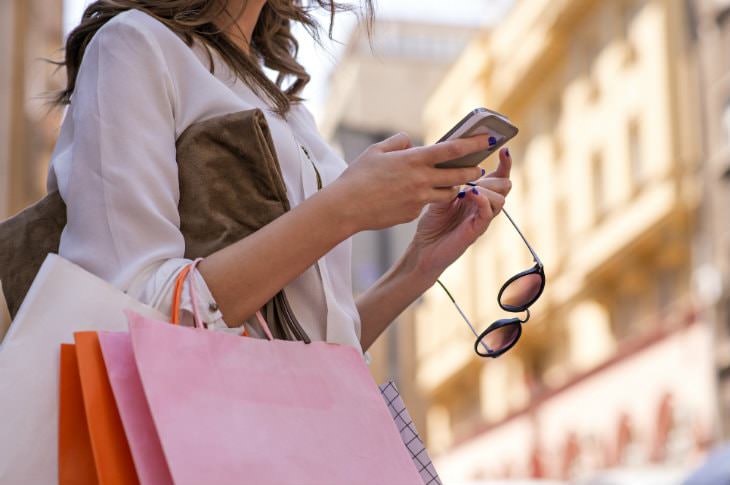 Woman with white, pink and orange shopping bags on her arm, holding her sunglasses, looking at her cell phone