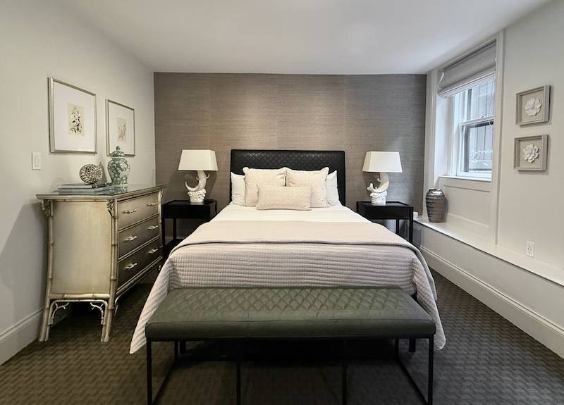 Bedroom with white walls, silver grass cloth wallpaper behind a black headboard. Dark green carpet and window onto garden.