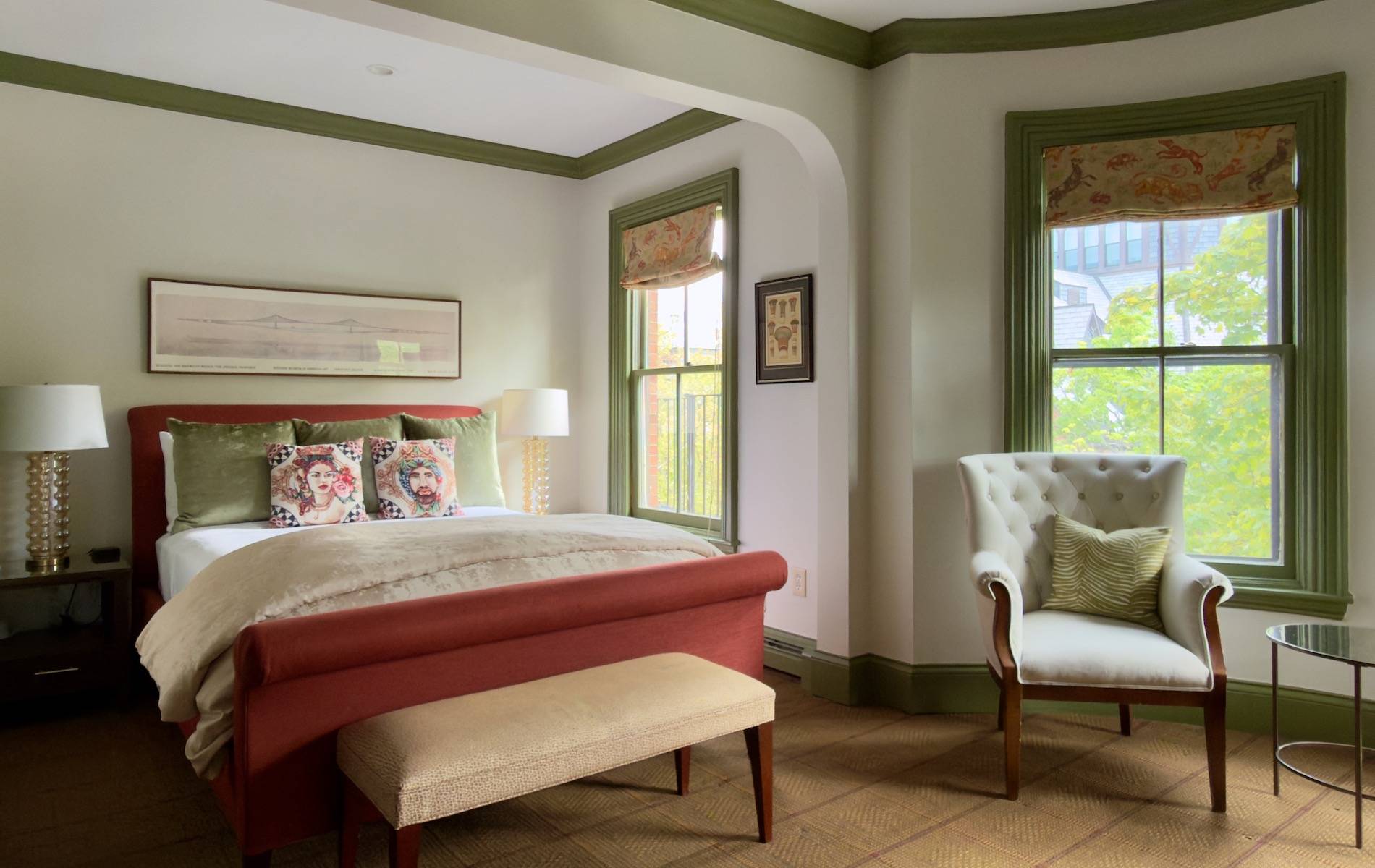 Hotel bedroom with a red upholstered bed, green window trim, two matching lamps, and decorative pillows in front of large windows.