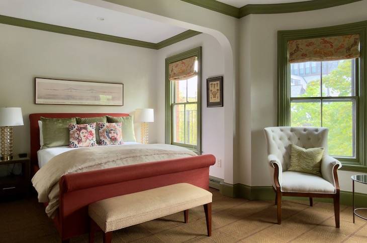 Stylish hotel bedroom with red upholstered bed, green accents, and a tufted white armchair by a window.