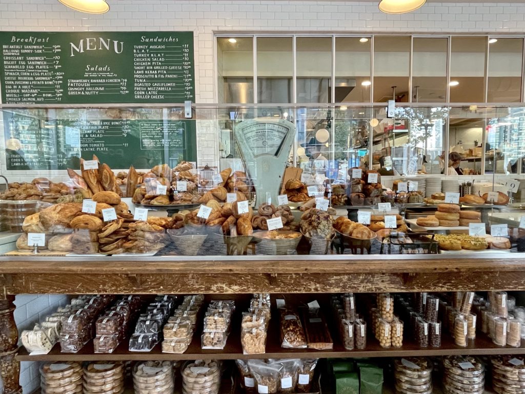 wooden table with glass partition containing pastries, croissants, scones, muffins on top, and rows of cookies biscotti and biscuits on shelf below green chalk board with white letters behind