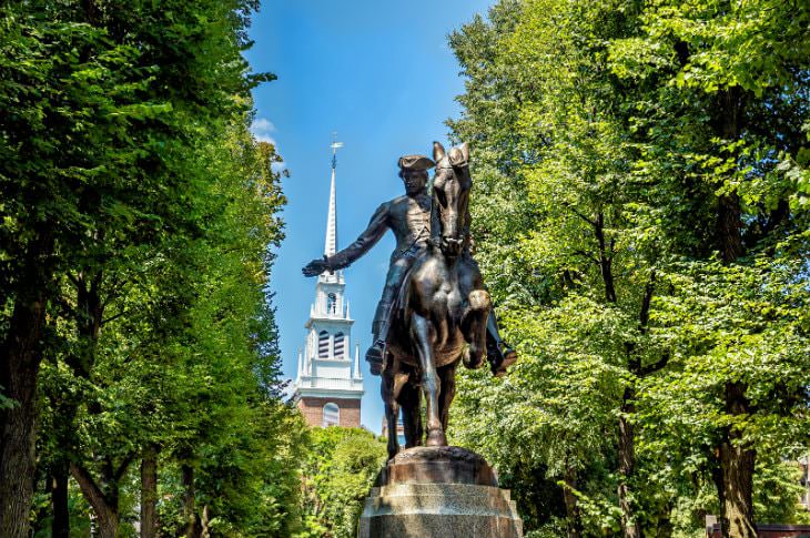 Bronze statue of Paul Revere surrounding by lush green trees with blue skies and a white steeple in background