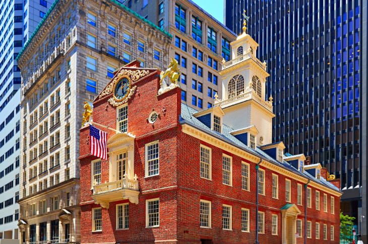 Small, red brick building with cream trim alongside taller stone and glass buildings