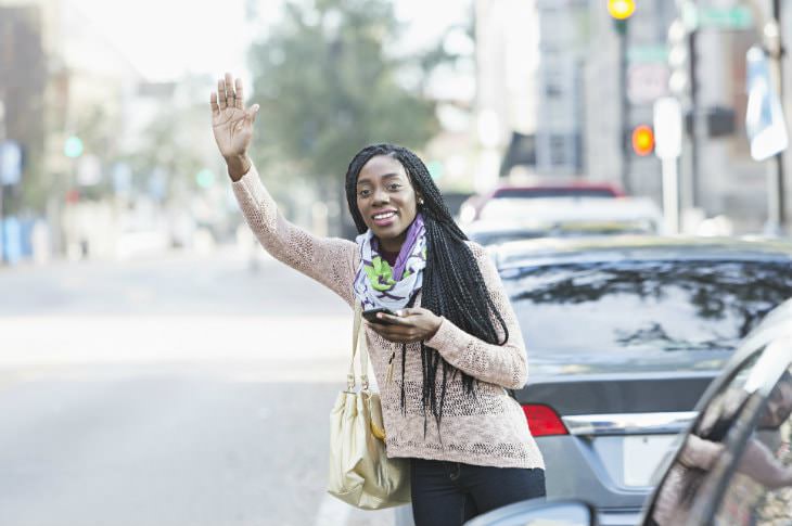 Woman wearing dark jeans and tan sweater waving toward a taxi while holding a cell phone